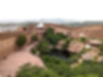 Historic fort wall in Jodhpur with steps and greenery. White temple with flag in the center. Waterhole surrounded by rocks and vegetation. Jodhpur the Blue City of India