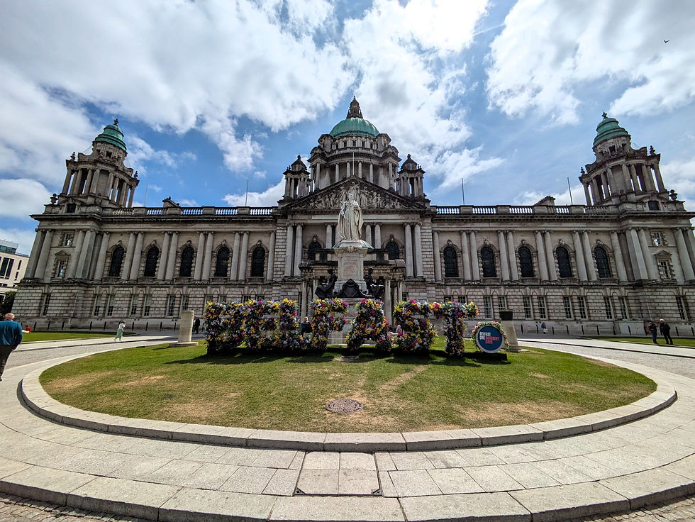 Belfast City Hall under a bright cloudy sky, with a floral BELFAST display and a statue in the foreground. The best things to do in Belfast City.