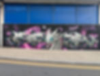 White dragon graffiti with teal eyes and pink cosmic swirls on a black wall beneath blue windows along a sidewalk. Street Art Murals of London