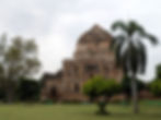 Ancient stone mausoleum with a large dome in a park, surrounded by green trees and grass. Overcast sky, serene mood. People strolling. Tourist places to visit in Delhi.