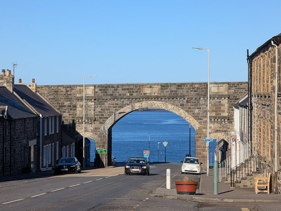 Street leading to a stone arch bridge with a view of the ocean. Two parked cars, brick buildings, and a clear blue sky in the background. Seafield Arms Cullen