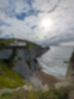 Coastal cliff with a white church and red roof under a cloudy sky. Sunlight reflects on the sea, creating a serene, picturesque scene. One of the best things to do in the basque country.