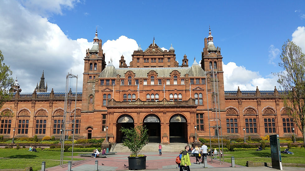 Kelvingrove Art Gallery in Glasgow, a red sandstone building with towers. People relax on the grass under a bright blue sky and clouds. Things to do in Glasgow Scotland.
