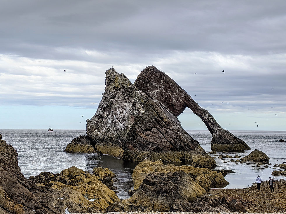 Rock arch in a coastal landscape with birds flying overhead. Two people walk on a rocky shore under cloudy skies. Boat visible in distance. Seafield Arms Cullen