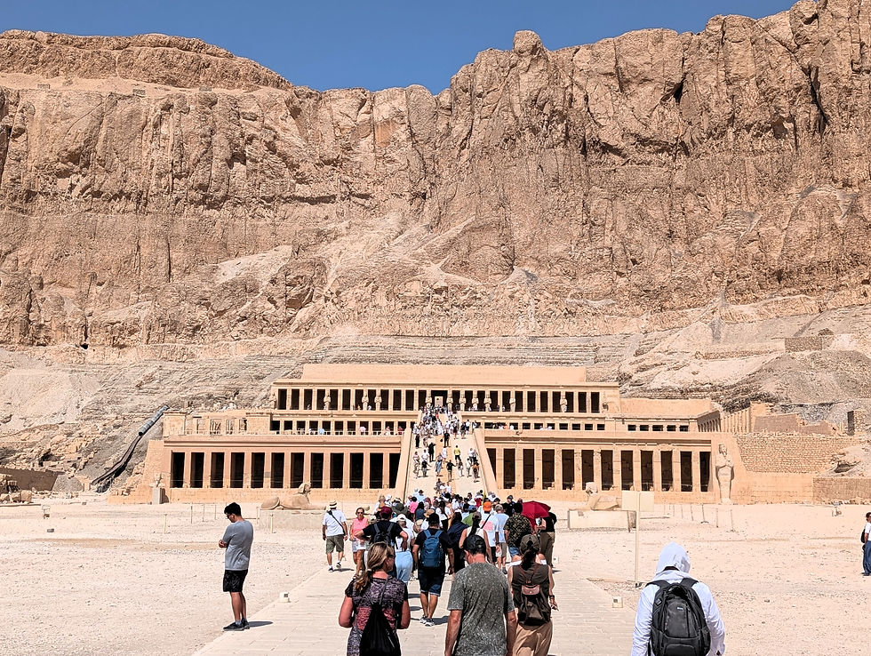 Tourists walk towards the ancient temple of Hatshepsut. The arid, rocky cliffs and clear blue sky form a dramatic backdrop. Things to Do in Luxor Egypt