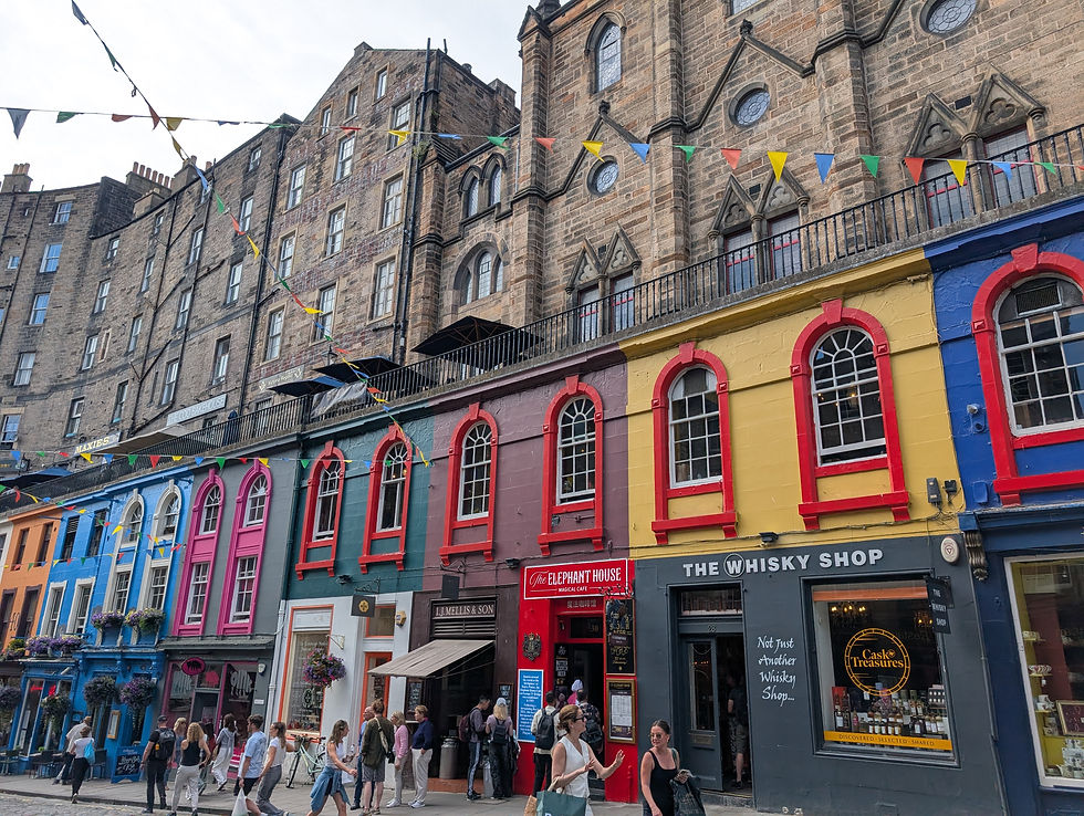 Colorful shops with red, blue, pink facades and a large stone building above. People walk on the street below bunting. Signs include The Whisky Shop. Harry Potter Places in Scotland