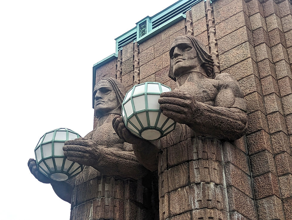 Two stone sculptures of muscular figures hold geometric lamps. They're part of a brick building with a detailed design, under an overcast sky. Art Nouveau and Art Deco cities around the world.