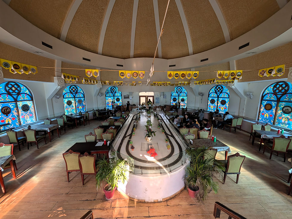 A bright restaurant with stained glass windows and wooden floors. People sit at tables around a central white model railway display. Tourist places to visit in Delhi