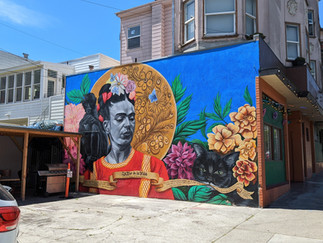 Street art murals on the side of a building depicting an image of Frida Khalo and a multi-coloured background.