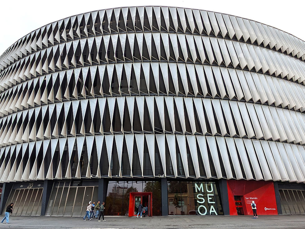 Modern building with geometric facade and large letters reading "MUSEOA." People walking nearby. Red accents, cloudy sky backdrop. One of the best things to do in Bilbao.