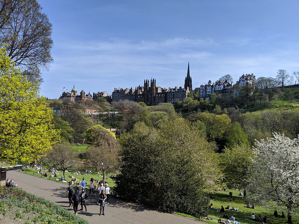 Park scene with people walking and sitting on grass. Historic buildings in the background under a clear blue sky. Lush green trees. Things to do in Edinburgh Scotland