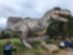 Large dinosaur statue among trees, kids taking photos, people seated on grass, cloudy sky. Relaxed and curious mood. Dinosaur days out in the UK.