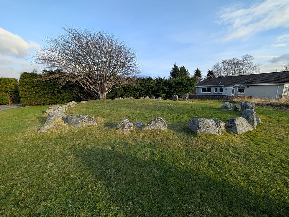 Stone circle on a grassy field with a leafless tree in the center. A house and evergreens in the background under a blue sky. Things to do at Aviemore in Scotland.
