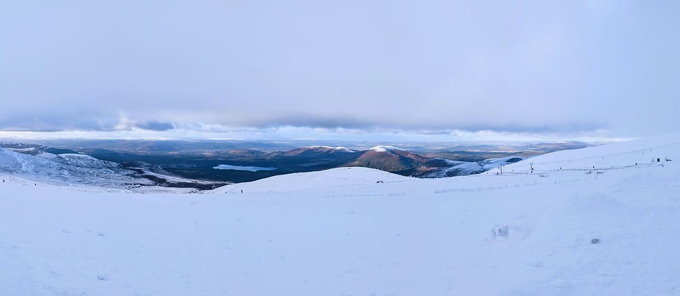 Snow-covered mountains under a cloudy sky with distant views of forests and a lake. A serene, winter landscape with soft blues and whites. Things to do at Aviemore in Scotland.
