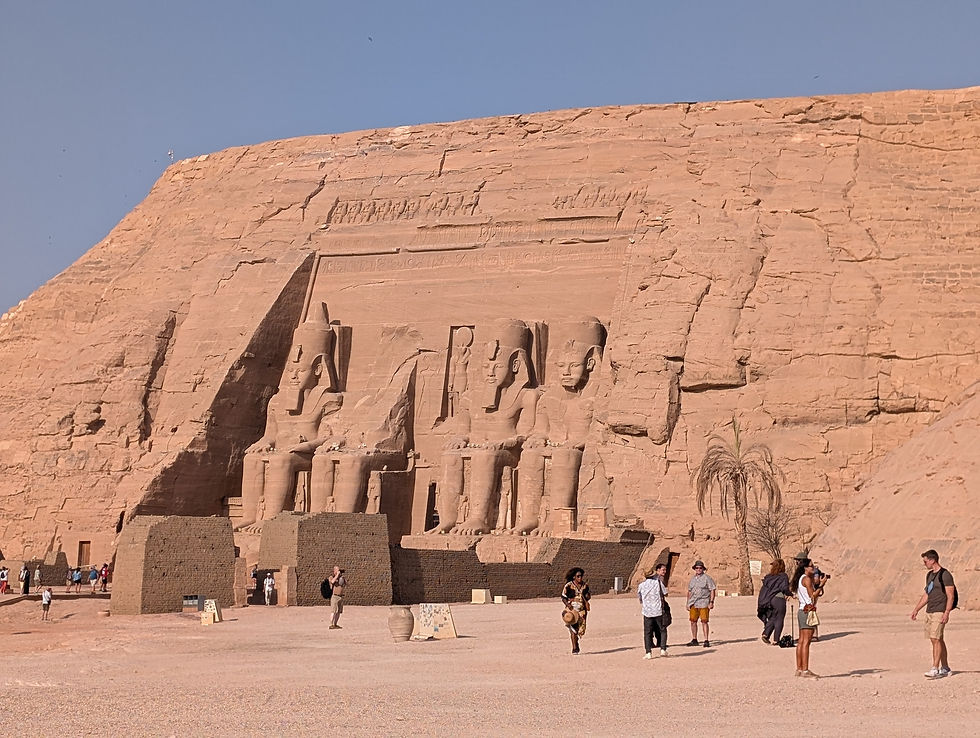 Tourists explore Abu Simbel temples with giant statues in a desert setting. Sunny day, sandy ground, clear blue sky. Ancient carvings visible. Luxor Aswan Nile Cruise