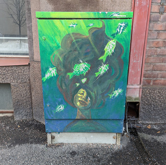 Street art murals painted on an electrical junction box on a city street depicting a woman with long hair under water, with fish swimming past and the water changing colour from green at the top to blue at the bottom.