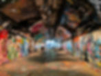 Graffiti-covered tunnel with arched brick ceiling, bright murals, and a few pedestrians walking toward the lit exit. Street Art Murals of London