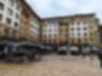 People sit under umbrellas at outdoor cafe tables in a town square with a fountain, surrounded by beige buildings with white-trimmed windows. Making this one of the best things to do in Bilbao.