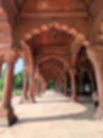 Red sandstone arches of historical architecture with people walking on the sunlit path. Lush green trees in the background. Tourist places to visit in Delhi.