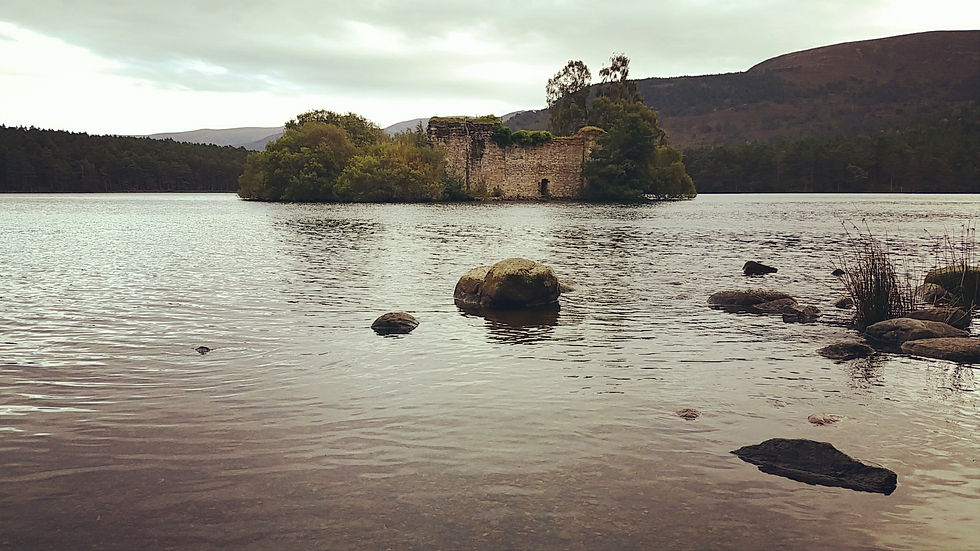 Ruins of a stone building on a small island in a serene lake, surrounded by trees. Overcast sky and scattered rocks in the foreground. Things to do at Aviemore in Scotland.