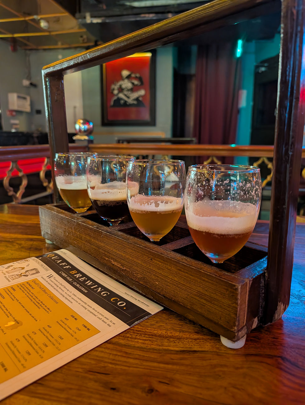 Four beer glasses in a wooden holder on a table with a Quaff Brewing Co. menu. Background has a red-framed art piece and soft lighting. Microbreweries in Gurgaon.