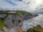 Coastal cliffs with a white building atop, overlooking waves crashing below. Lush green hills in the background under a cloudy sky. One of the best things to do in the basque country.