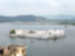 White palace floating on a lake with green hills in the background. Birds are in the sky, creating a serene scene under a cloudy sky.things to visit in Udaipur