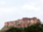 A majestic ancient fort stands atop a hill with lush greenery, under a cloudy sky, showcasing beige stone walls and detailed architecture. Jodhpur the Blue City of India