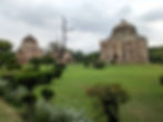 Historic stone mausoleums with domes in a lush green garden. Cloudy sky, trimmed bushes, and small flowers create a serene atmosphere. Tourist places to visit in Delhi.