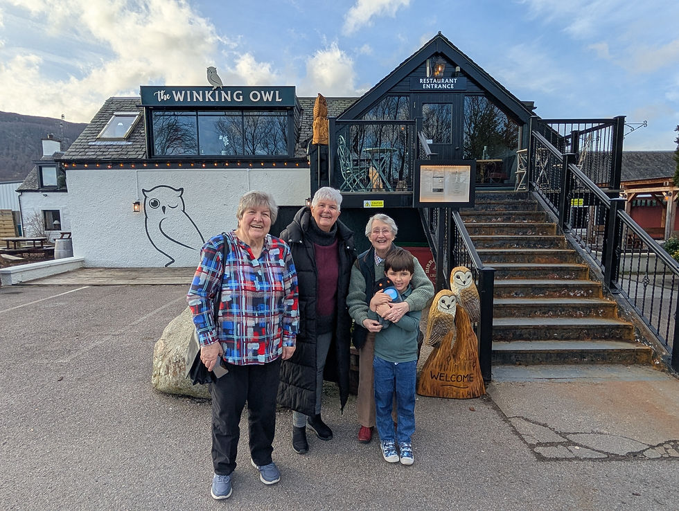 A group of three adults and a child smile outside "The Winking Owl" restaurant. The building features owl art and a wooden owl sign. Things to do at Aviemore in Scotland.