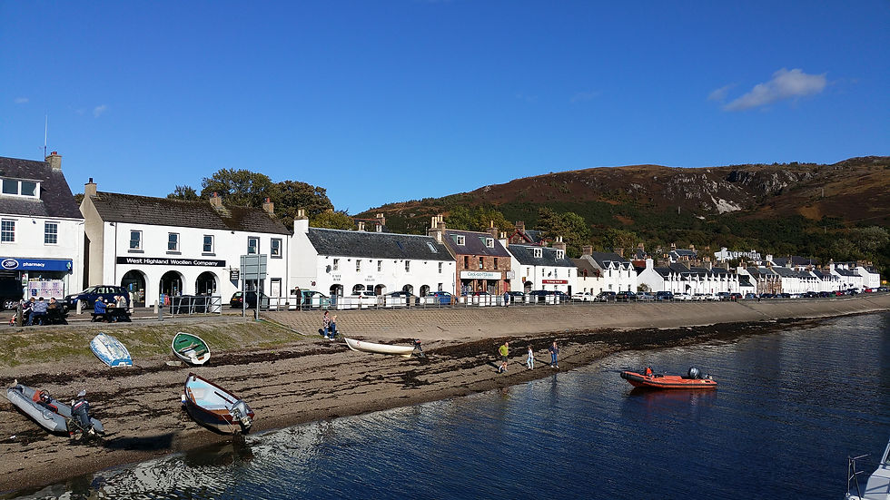 Sunny harbor village with white shops and boats on a rocky shore below a hillside, including West Highland Woollen Company. NC 500 Route - Scotland's Route 66