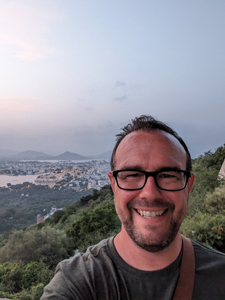 A selfie of a man taken from a hilltop, with the city of Udaipur below in the background, making it one of the attractions you should visit in Udaipur, India