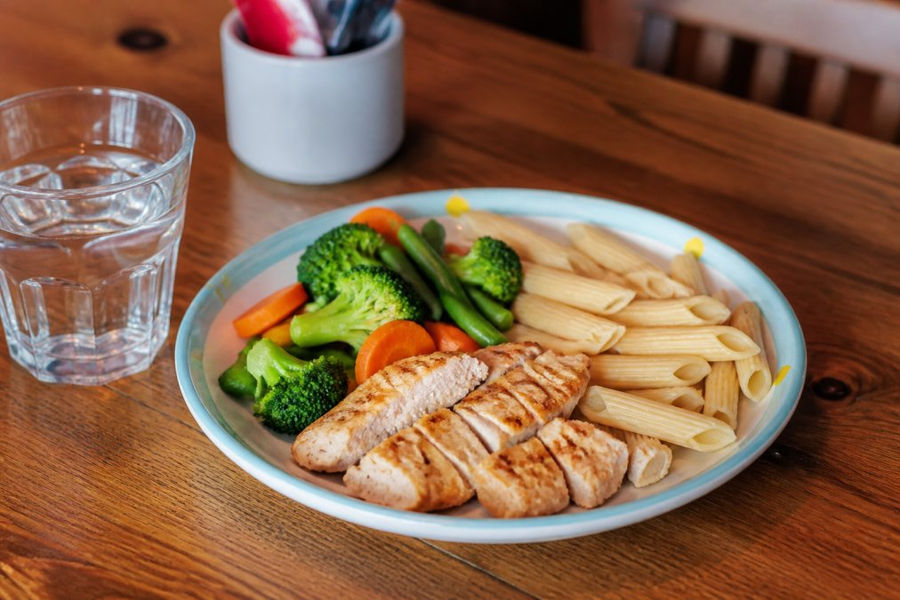 A colourful plate of child-sized portions featuring grilled chicken, steamed vegetables, and whole grain pasta on a wooden restaurant table, with a small glass of water beside it