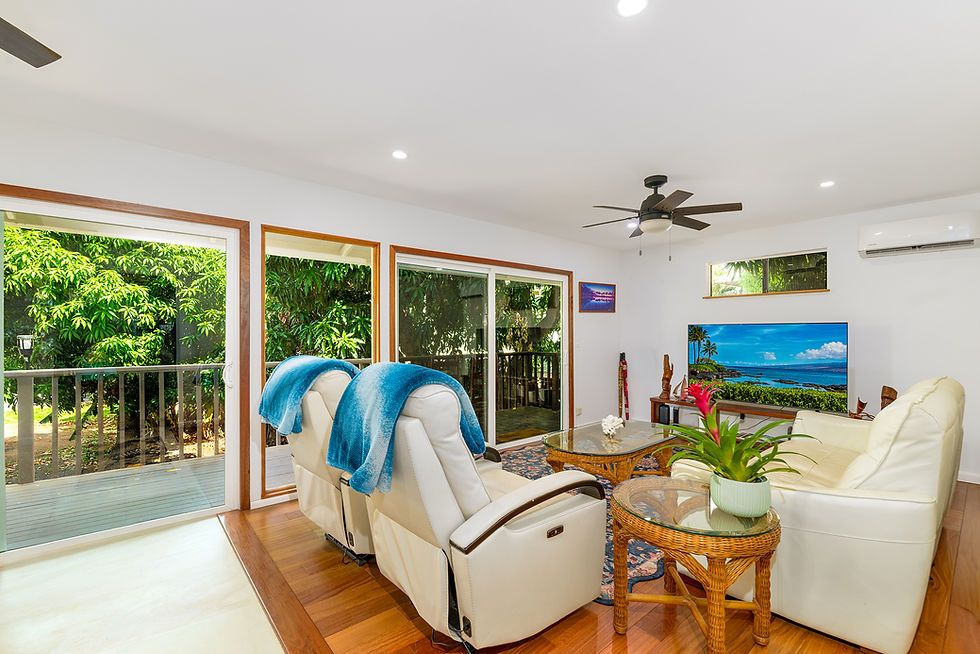 Bright tropical living room with white leather seating, blue throws, TV showing ocean view, and large windows to lush greenery.