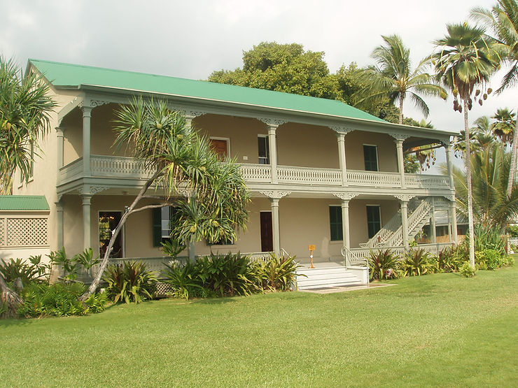 Two-story tropical house with green roof, white porch railings, palm trees, and a manicured lawn under a bright sky.