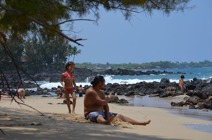 People relax and play on a rocky tropical beach with waves, palm trees, and a child in a pink bikini; sunny, carefree mood.