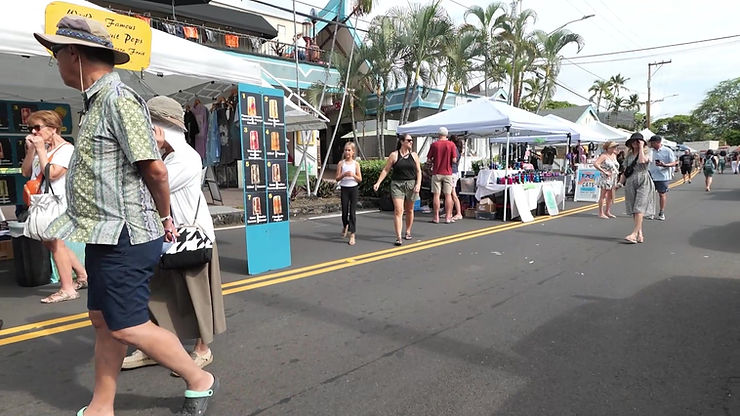 People browse white tents at a palm-lined street market, with a popsicle menu sign and casual shoppers on the road.