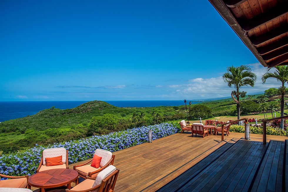 Ocean view from a wooden deck with chairs, tables, and vibrant flowers. Green hills and a bright blue sky in the background create a serene scene.