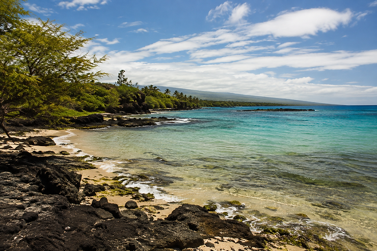 Rocky tropical beach with clear turquoise water, green trees, and a blue sky with wispy clouds along the coast.