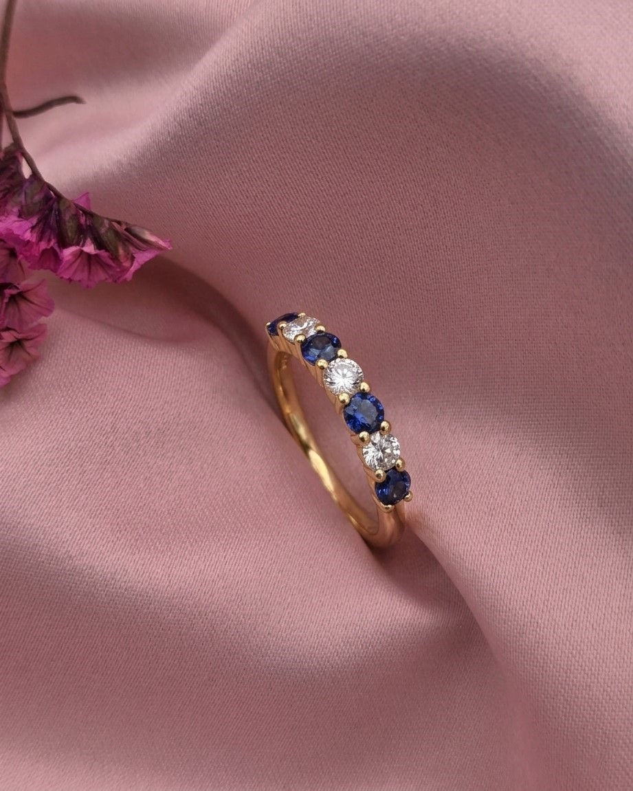 Anelem ouro amarelo com safiras e diamantes pousado sobre um tecido de cetim rosa com flores secas ao fundo.