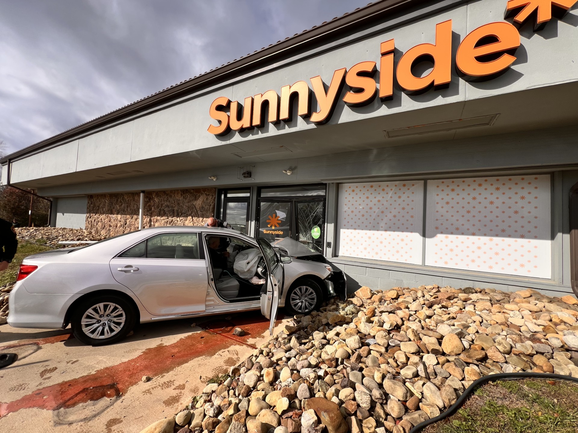 Silver car crashed into "Sunnyside" storefront. Deployed airbags visible. Overcast sky, river rocks in foreground, and orange lettering.