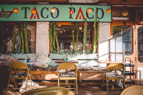 The Taco Paco Restaurant terrace lid up by the sun with tables, chairs and cosy benches. On the top you can see the Taco Paco logo and some cactus plants.