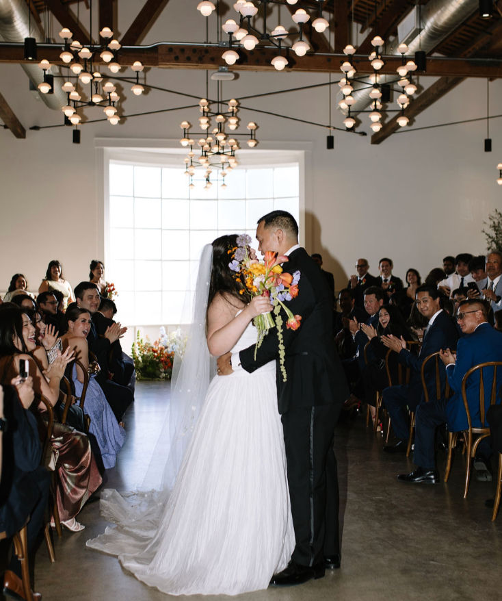 Luxury Southern California wedding ceremony with the bride and groom sharing a joyful kiss as loved ones applaud beneath modern chandeliers, highlighting timeless indoor wedding design.