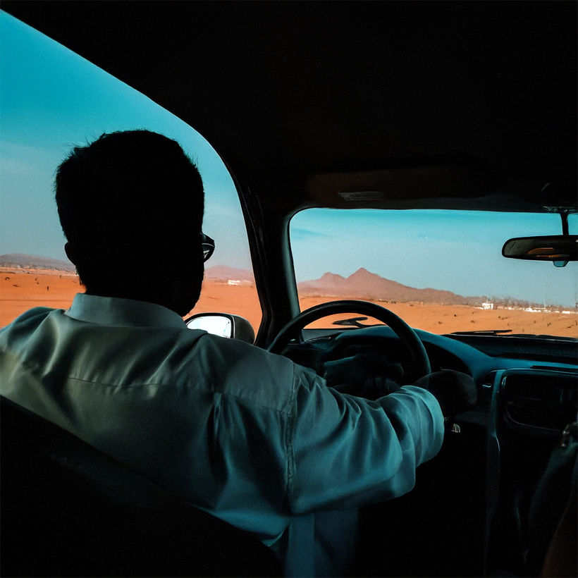 Person in white shirt driving a car through a desert landscape with distant mountains, under a clear blue sky. Mood is calm and focused.