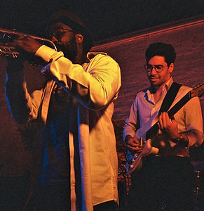 A live music performance featuring a close-up of a trumpet player in the foreground and a guitarist in the background