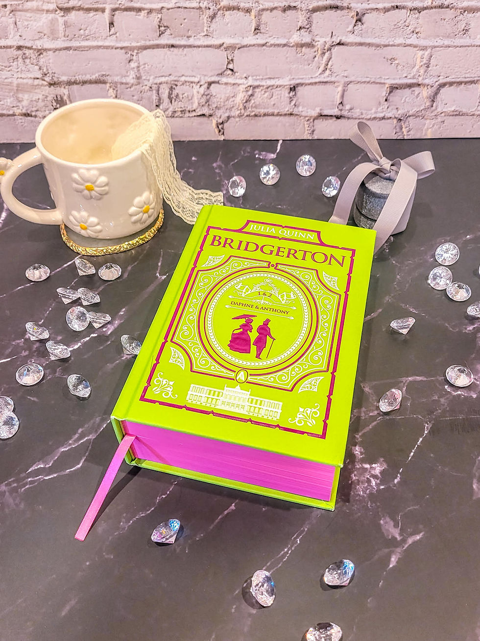 A bright green "Bridgerton" book with pink edges on a marble surface, surrounded by crystals, next to a daisy mug with lace and a ribboned jar.