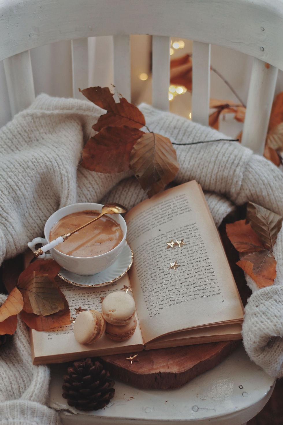Cozy autumn still life on a white chair: open book, coffee cup with gold spoon, macarons, dried leaves, and a pine cone.