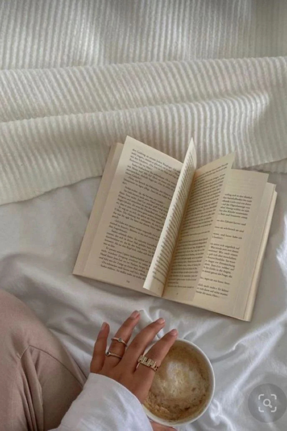 Hand holding a coffee cup beside an open book on a white textured blanket. The scene is calm and cozy, with neutral tones.