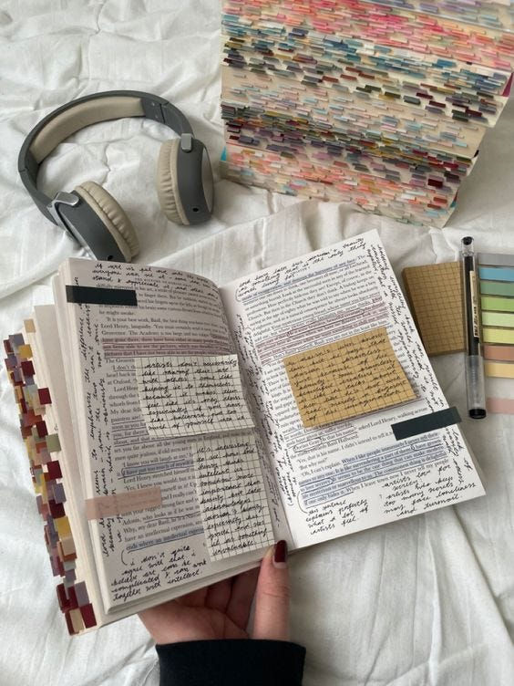 Hand holding an annotated open journal on a white bed, surrounded by headphones, a color-tabbed book stack, and a pen.