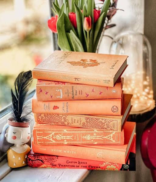 Stack of orange vintage books beside red tulips and a glowing lamp, with a small feathered figurine on a windowsill.